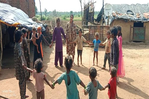 ग्रामीण बस्ती के खुले स्थान पर बच्चे और महिलाएं हाथ पकड़कर घेरा बनाए खड़े हैं
